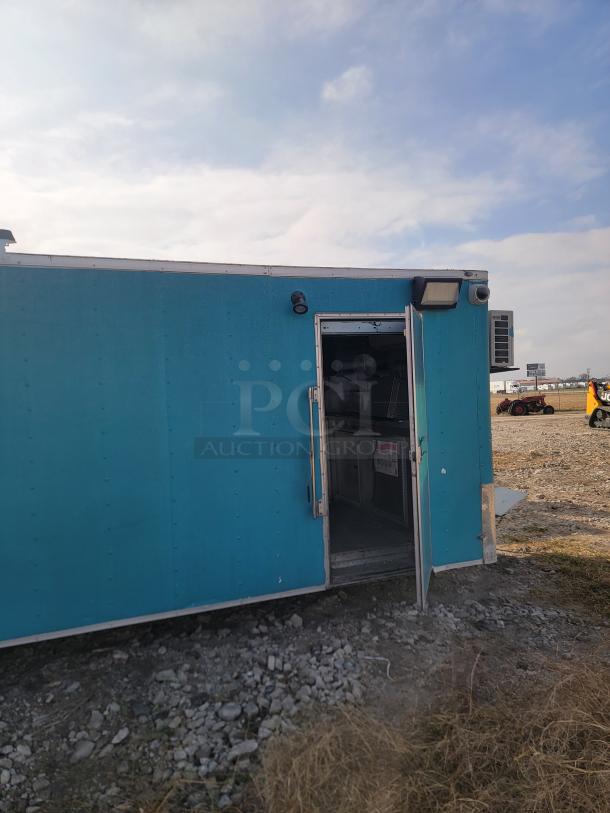 Blue utility trailer with an open door, showing interior workspace. Exterior features lighting and air conditioning unit.
