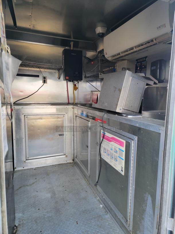 Interior of a commercial kitchen truck with stainless steel counters, shelving, and appliances. Ventus air conditioning unit mounted on the wall. Ceiling-mounted camera and wall-mounted heater visible. Clean and well-maintained.