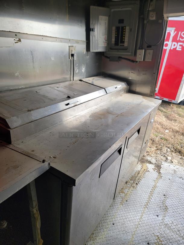 Commercial stainless steel prep table with storage compartments in a food truck. Some surface wear, electrical panel nearby.