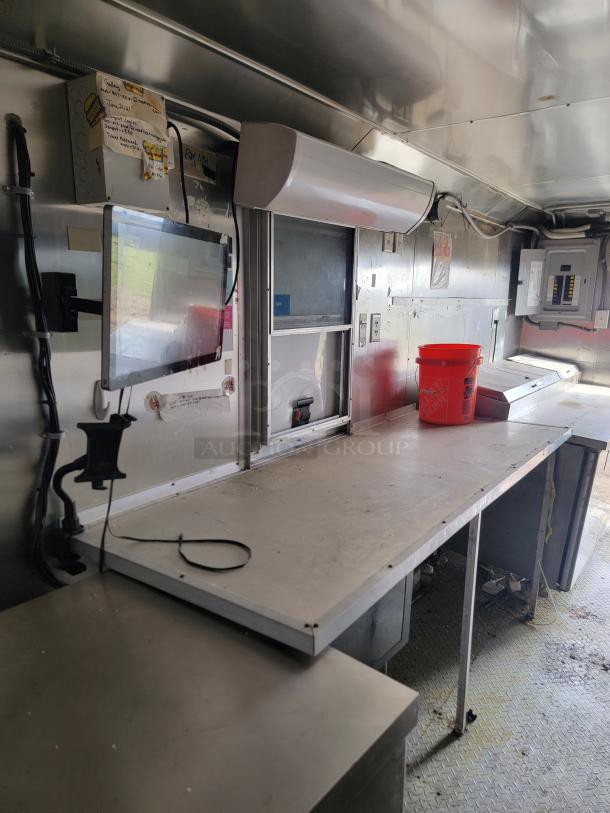 Inside of a stainless steel food truck with work surfaces, electrical sockets, a wall-mounted monitor, and a red bucket.
