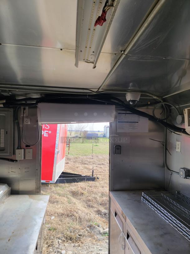 Interior view of a metal trailer with stainless steel surfaces, shelving, and electrical outlets. Visible outdoor field and red trailer.