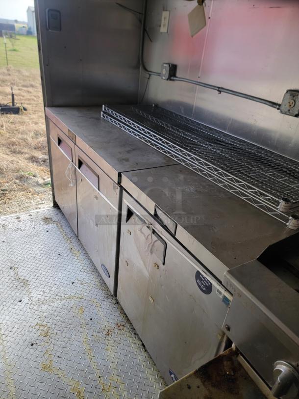 Stainless steel industrial kitchen counter with storage, showing signs of wear, featuring a wire rack and electrical outlets.