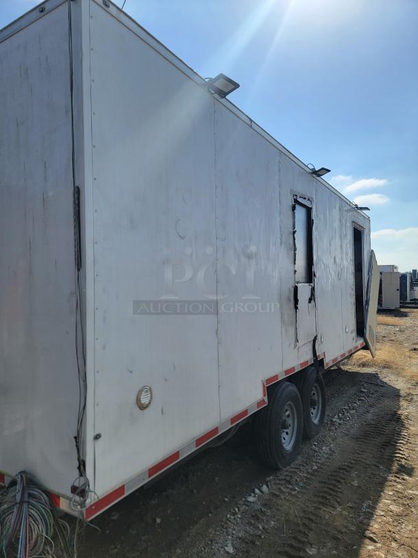 White mobile trailer on double axles, visible damage on panels, exterior wiring, and rear door partially open. Appears used.