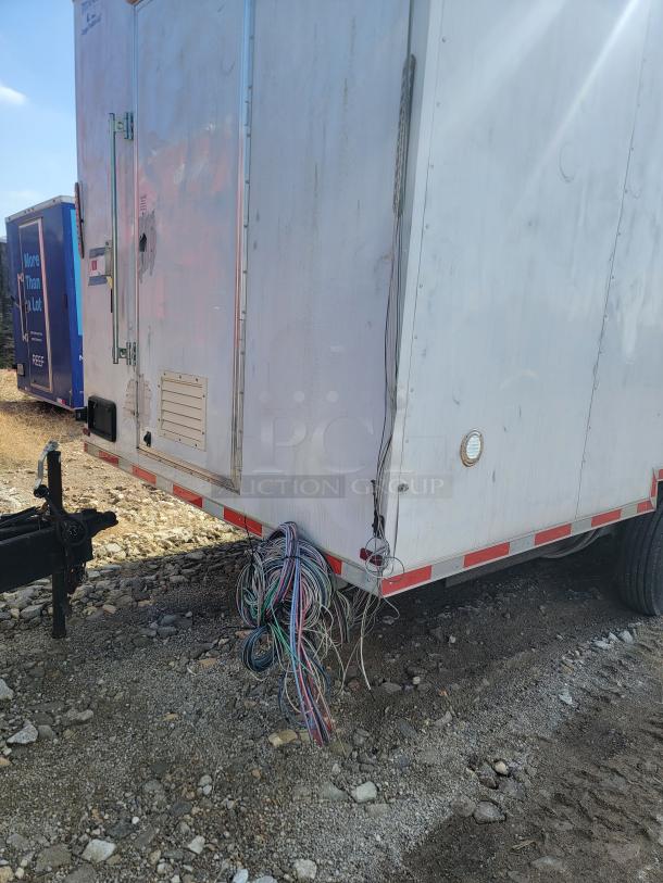 White utility trailer with wire bundles, noticeable wear, and front hitch, parked on gravel. Marks and labels visible but unreadable.