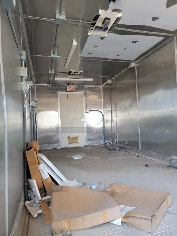 Interior view of an empty metal trailer with electrical wiring, loose panels, and debris on the floor. Exit sign above rear door.
