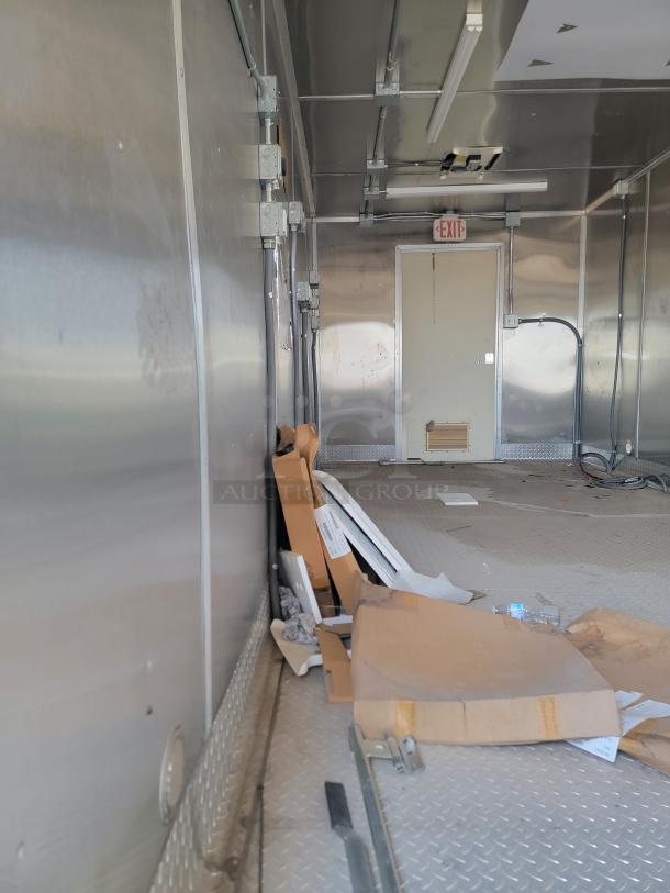 Interior of a metal transport trailer with exposed electrical wiring, ceiling lights, and an exit door. Assorted boxes and debris on floor.