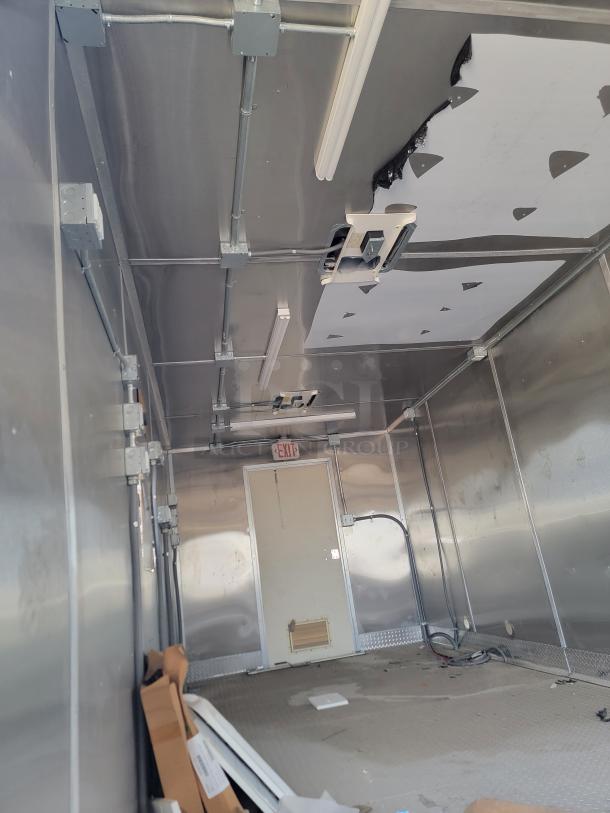 Interior view of a metallic industrial trailer with visible wiring and damaged ceiling panels. Exit door marked at the rear.