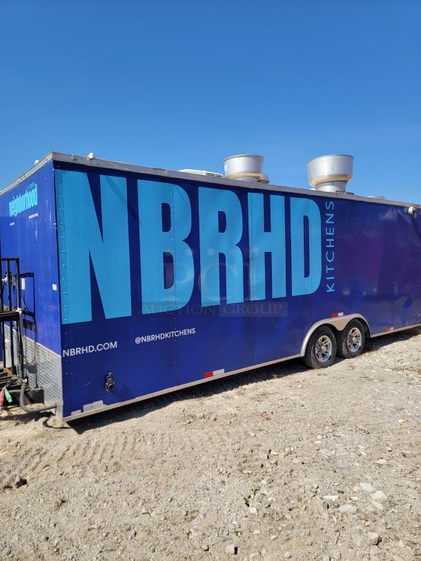 Blue mobile kitchen trailer labeled "NBRHD Kitchens" with ventilation units on the roof, seen on a gravel lot.