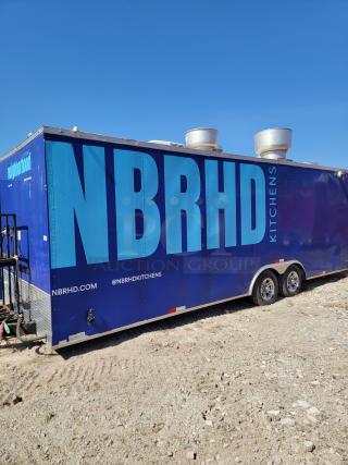 Blue mobile kitchen trailer labeled "NBRHD Kitchens" with ventilation units on the roof, seen on a gravel lot.