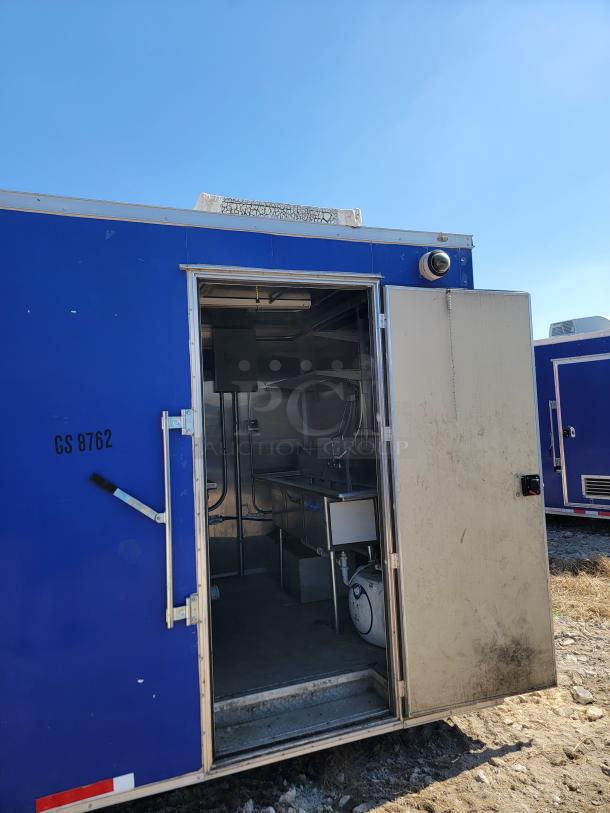 Blue mobile trailer with open door, revealing stainless steel interior kitchen setup. Visible marking: "GS 8762." Weathered exterior.