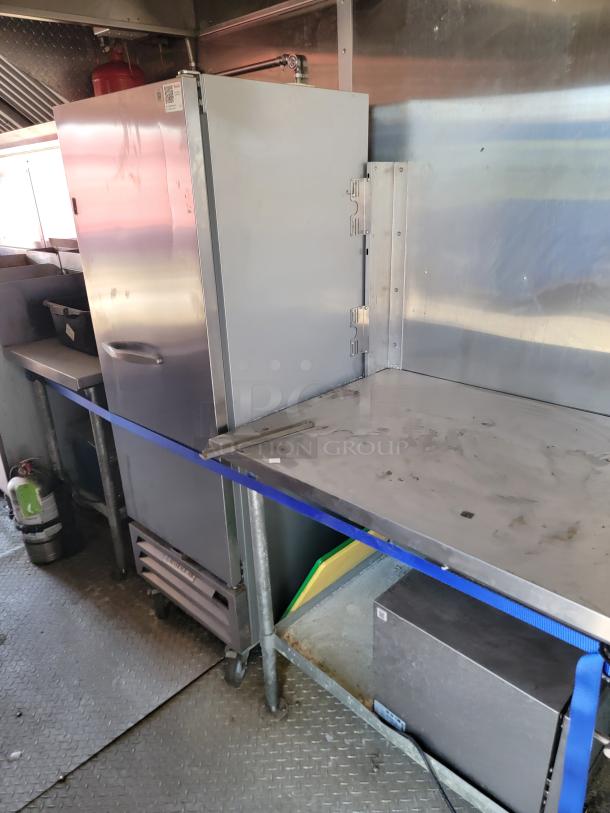 Commercial stainless steel refrigerator in a kitchen, with visible wear. Includes shelves and blue strap for stability.
