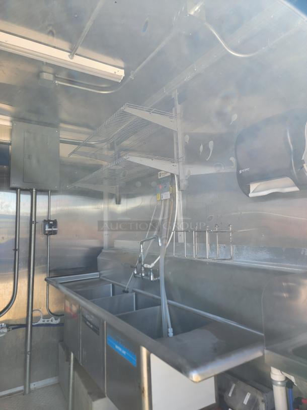Commercial kitchen trailer interior with stainless steel triple sink, overhead shelving, and paper towel dispenser. Appears in good condition.