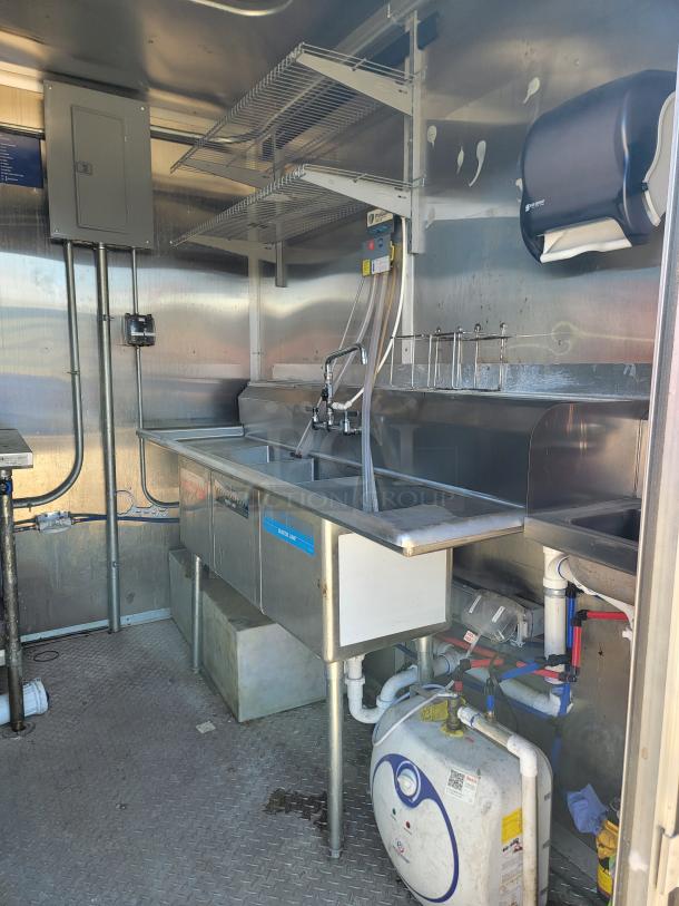 Commercial stainless steel kitchen setup with triple sink, wall-mounted shelves, plumbing fixtures, and paper towel dispenser.