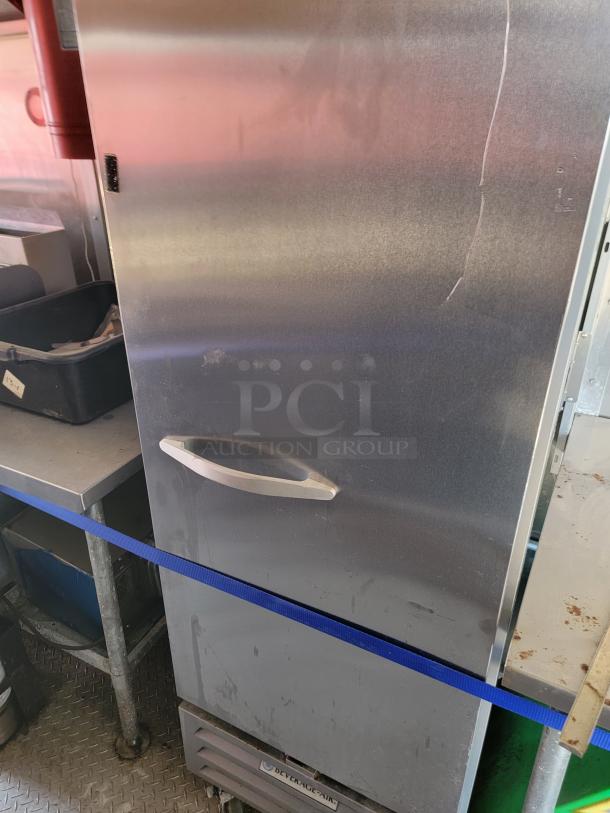 Stainless steel commercial refrigerator with visible surface wear and a blue strap securing the door. Suitable for restaurant use.