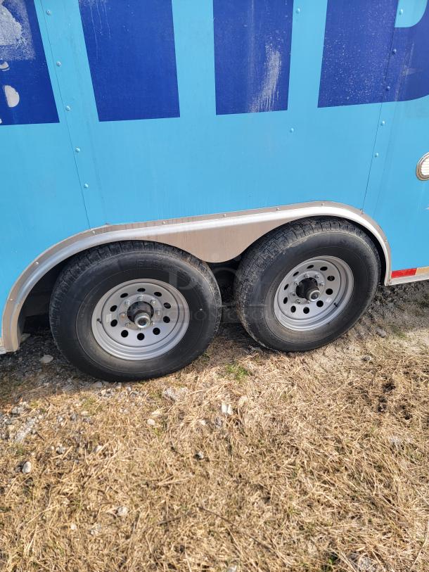 Blue trailer's dual wheels on dry grass, showing silver rims and black tires. Side panel is worn with slight dirt marks.