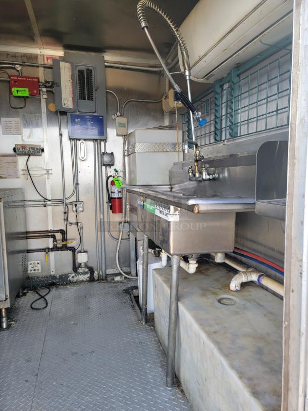 Industrial kitchen interior with stainless steel sink and sprayer, electrical panel, and fire extinguisher; signs of wear.