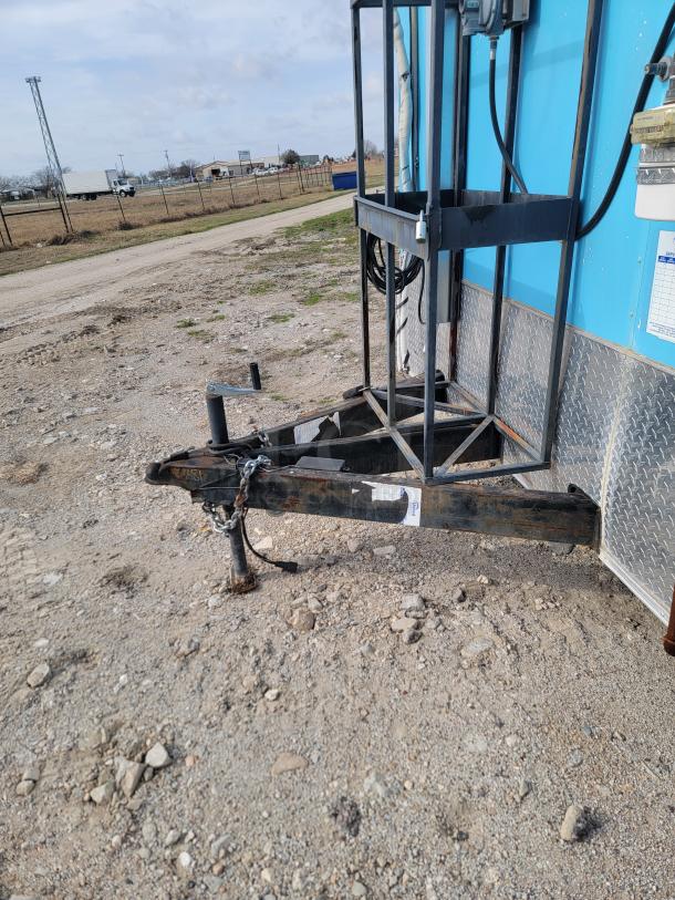 Close-up of a trailer hitch on a blue mobile unit with metal framing. Chain and hook attached. Rough, gravel ground.