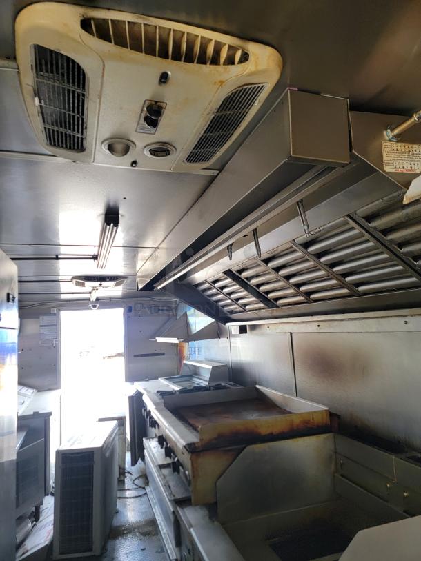 Commercial kitchen interior with vent hoods, gas grill, and ceiling air vent. Slight wear, functional workspace setup.