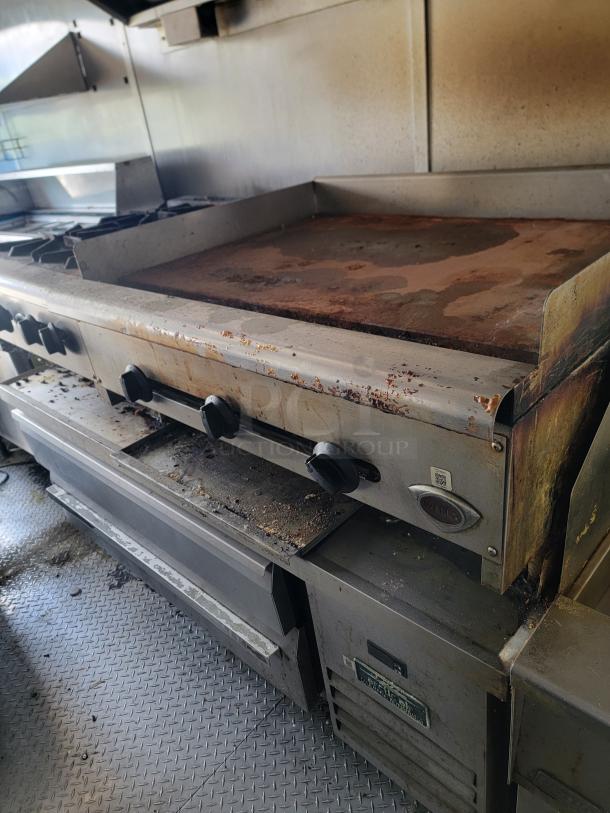 Commercial griddle with stove, rust on surface, three control knobs. Marks of use on metal chassis, mounted on textured floor.