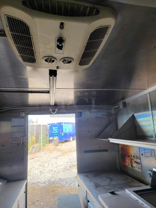Interior of a commercial kitchen space with stainless steel surfaces and a ceiling air vent. Outdoor view through open door.