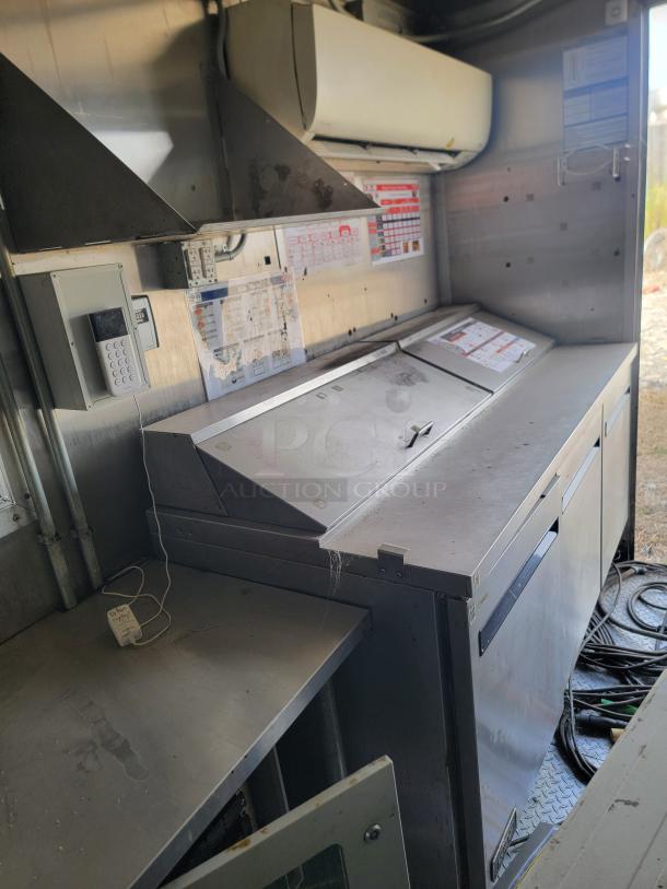 Metallic industrial kitchen equipment, featuring a stainless steel prep station with covered compartments and visible vent hood. Appears used, with control panels and wiring visible. Some instruction labels are affixed to the walls.