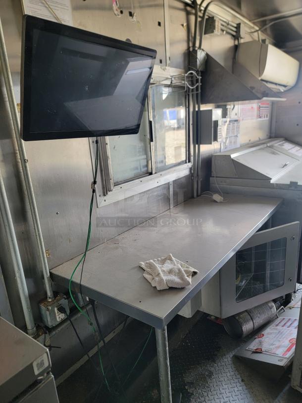 Commercial kitchen counter with stainless steel surface, wall-mounted monitor, and nearby window in used condition.