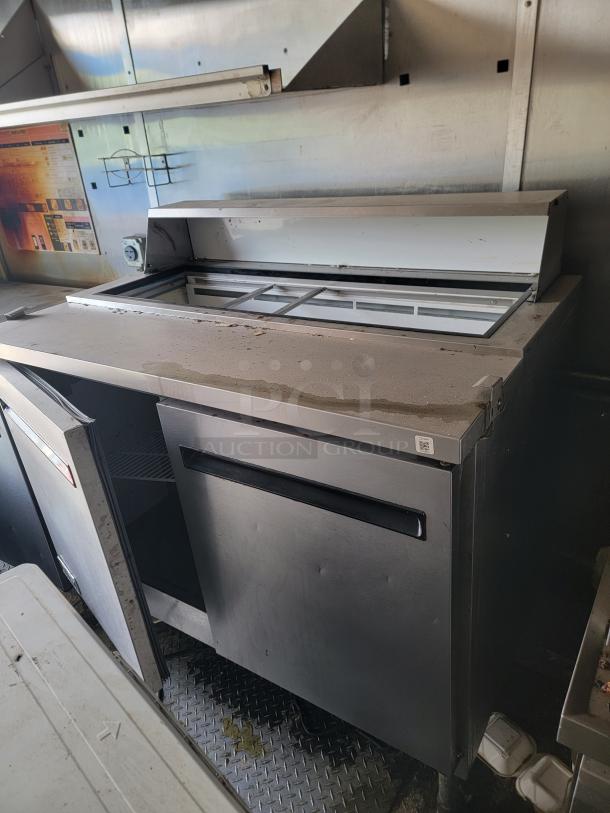 Industrial stainless steel refrigeration unit, shows signs of wear, open door, interior shelf, utility in commercial settings.