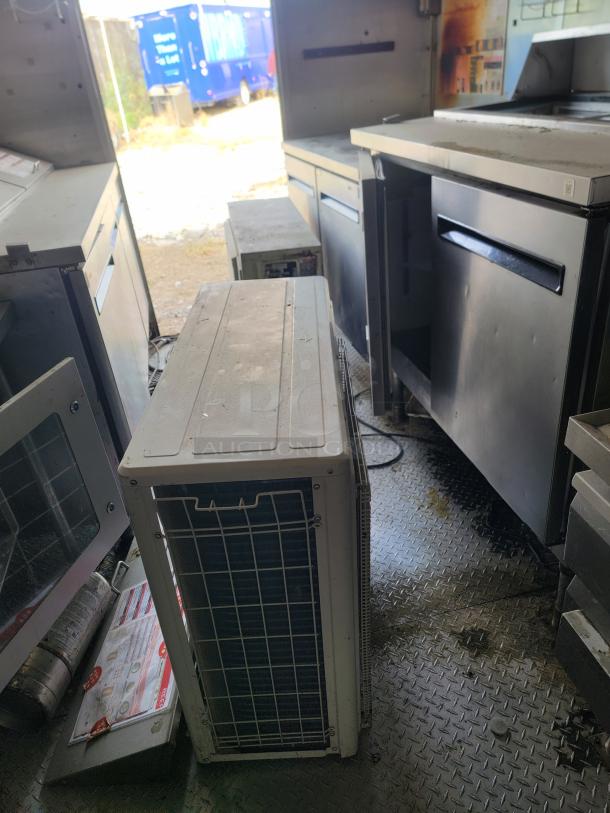 Industrial kitchen equipment inside a trailer, featuring stainless steel counters and cabinets, with an air conditioning unit.