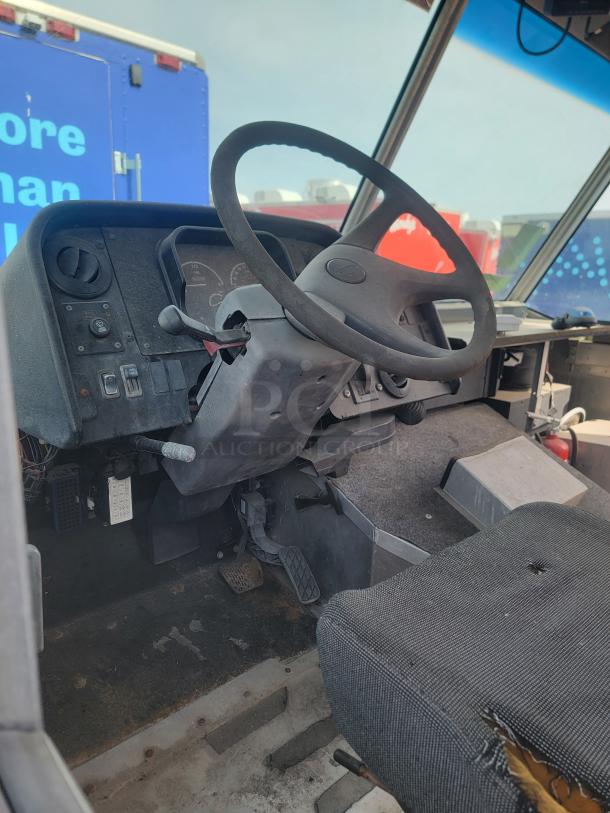 Bus driver cockpit, worn fabric seat, visible steering wheel, pedals, and dashboard. Some wear and tear evident.