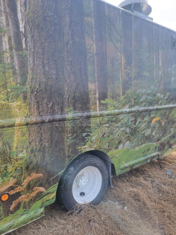 Side view of a vehicle with a forest-themed wrap, showing lush greenery and tree imagery. White wheel visible, parked on dry grass.
