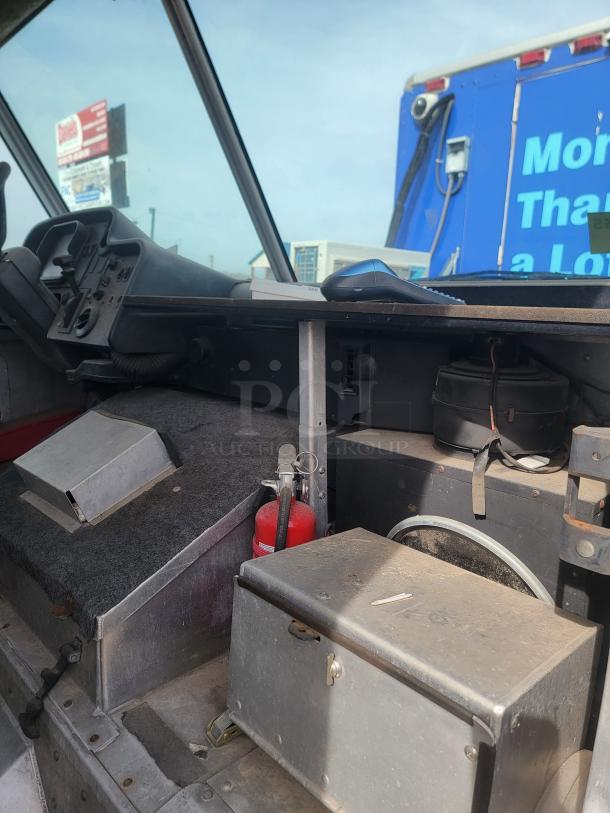 Interior of a vehicle showing a dashboard, steering components, and storage compartments. A fire extinguisher and metal box are visible, indicating emergency readiness and storage function. Dust and wear suggest usage and age.