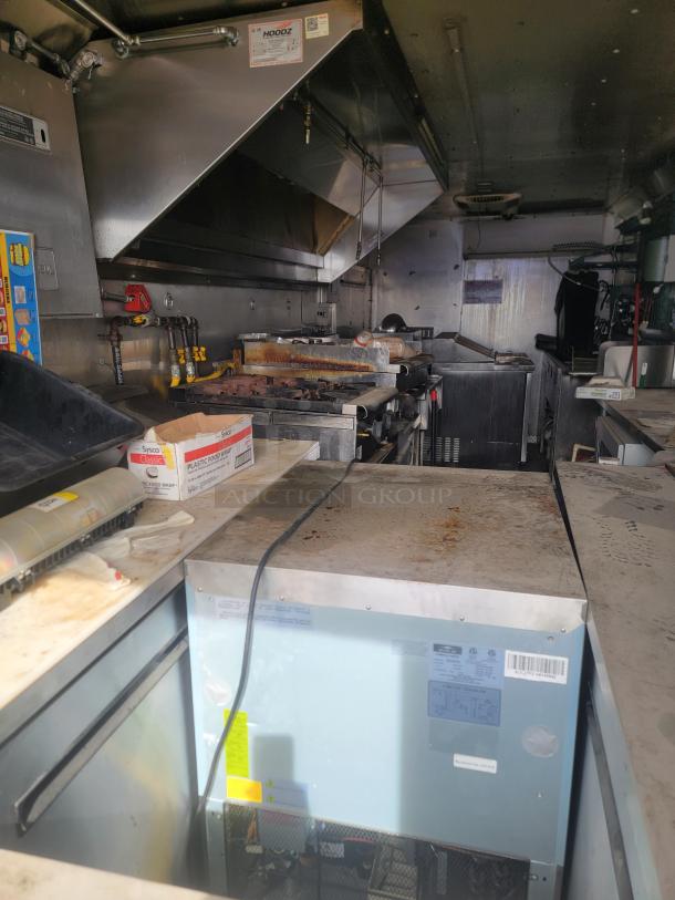 Interior of a commercial kitchen with stainless steel appliances, hoods, a stove, and equipment. Surfaces show signs of use.