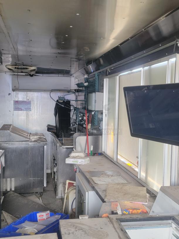 Interior of a used mobile kitchen or food truck, featuring industrial stainless steel equipment in fair condition, with visible cleaning supplies, fryers, and a mounted screen. Dust and clutter present.