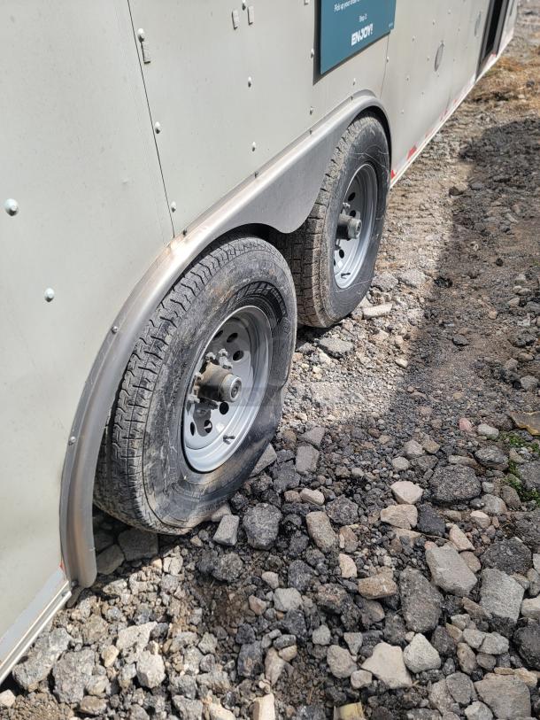 Dual-axle trailer with one flat tire, gray metal body, slight wear. Visible markings and rivets on the side. Gravel ground.