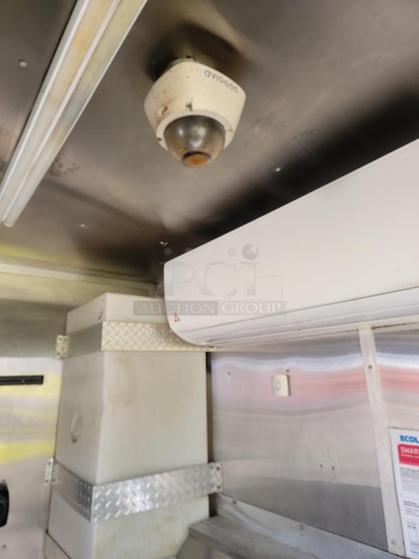 Avigilon security camera mounted on ceiling inside a vehicle. Visible air conditioning unit and storage container.