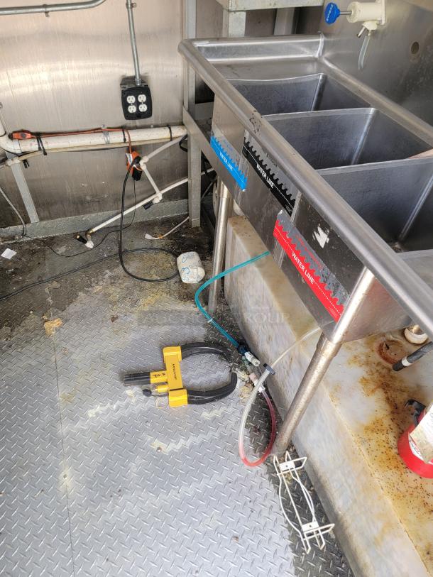 Commercial kitchen sink with triple basins, labeled water lines, visible wiring, valved pipes, and a yellow clamp tool on the floor.