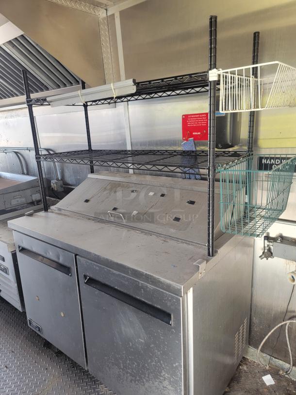 Commercial stainless steel prep station with shelving, baskets, and storage compartment. Used condition with visible markings. Ideal for food service.
