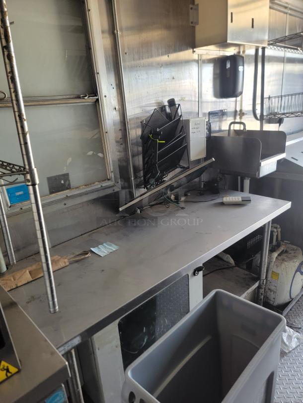 Commercial kitchen workspace with stainless steel countertop, shelving, paper towel dispenser, and organizer. Visible wear.