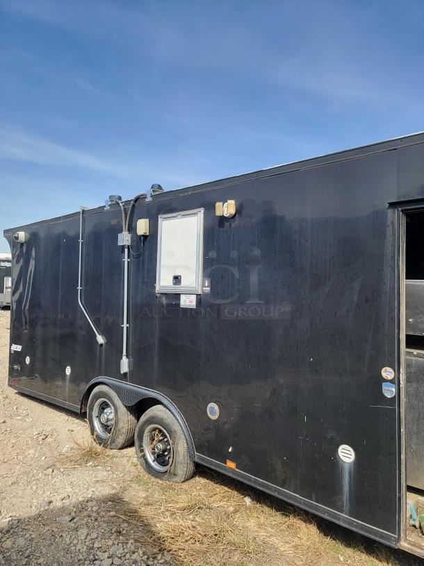 Black utility trailer with dual axles, side door, and exterior power outlets. Slightly worn exterior; parked outdoors on rocky ground.