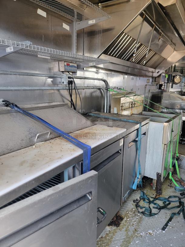 Commercial kitchen setup with stainless steel appliances and shelving. Features include refrigeration units and ventilation hood. Visible wear and straps securing equipment.