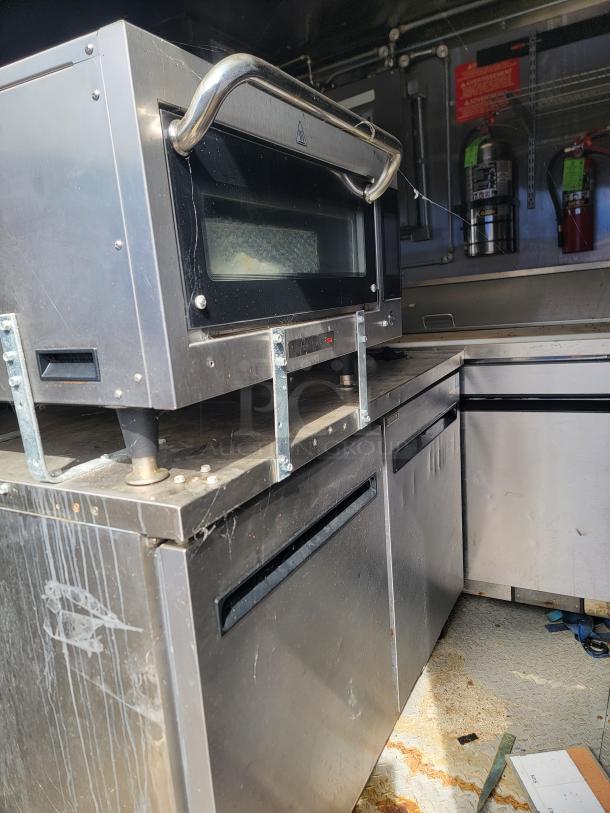 Stainless steel commercial oven with handle, mounted on metal brackets, showing signs of wear. Fir extenders in background.