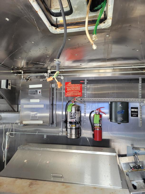 Industrial kitchen interior with mounted fire extinguishers, water pipes, and a paper towel dispenser. Stainless steel surfaces.