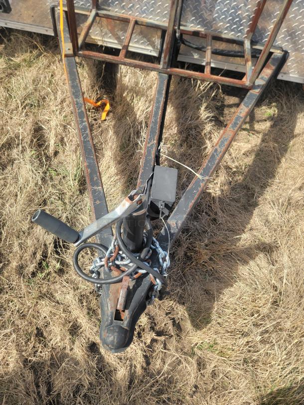 Trailer hitch with attached chains, lever, and wiring, in worn condition on grass. Metal frame visible with rust spots.