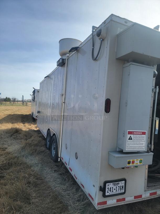 White utility trailer with dual axles in a grassy field, equipped with various control panels and safety warnings. Texas license plate visible.