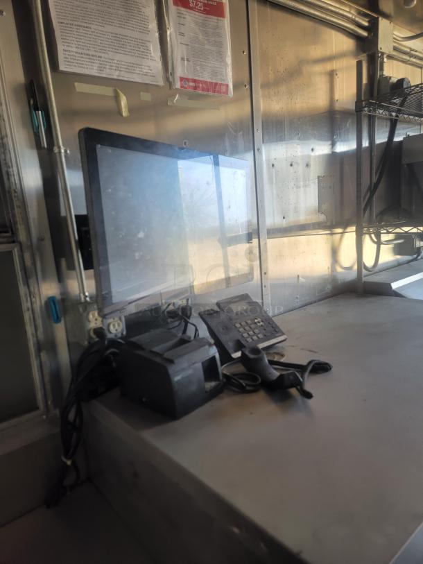Industrial workstation with monitor, phone, printer, and handheld scanner on a steel countertop. Wall displays labor notices.