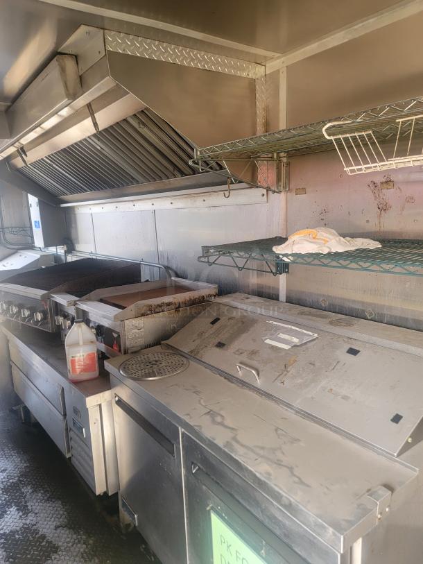 Commercial kitchen equipment with stainless steel prep table, grill, hood vent, and shelves. Visible signs of use.