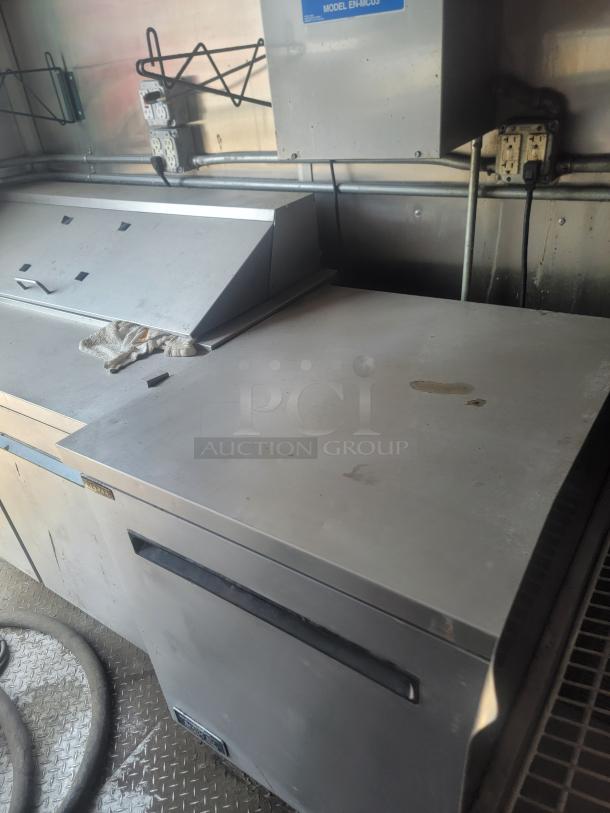 Commercial stainless steel prep table with upper compartment, electrical outlets on wall, and metal shelving above. Moderate wear.