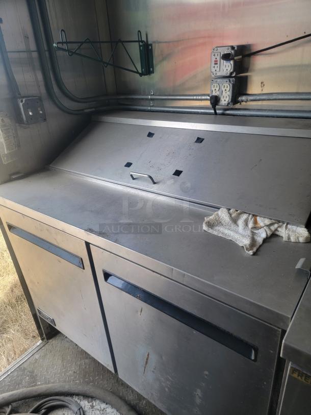 Stainless steel commercial refrigerator unit with top lid, showing outlets and wire shelving. Some visible wear and minor stains.