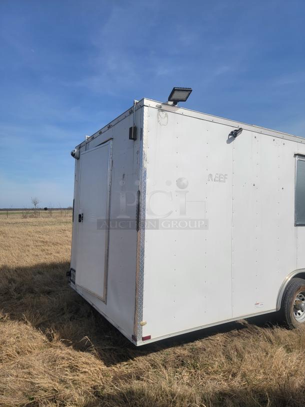 White utility trailer with rear door and side handle, minimal wear. Features mounted floodlights and visible "REEF" marking.