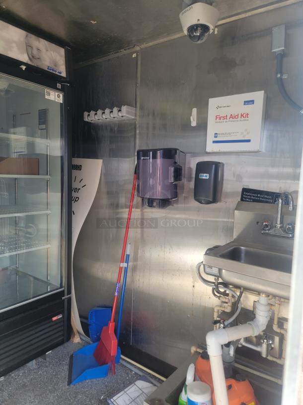 Industrial metal utility area with a Turbo Air fridge, sink, first aid kit, cleaning tools, and dispensers.